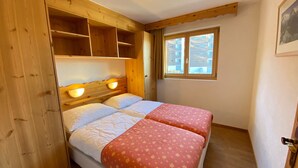 A bedroom with a wooden headboard, two beds with floral bedding, a window with a view of buildings, and a door. - Pracondu 1 101 - OUTDOOR & FUN  apartment 8 pers (Haute-Nendaz)