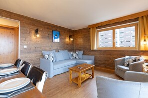 Een moderne woonkamer met een houten wand, een blauwe bank, een houten salontafel en een eethoek met borden en bestek.