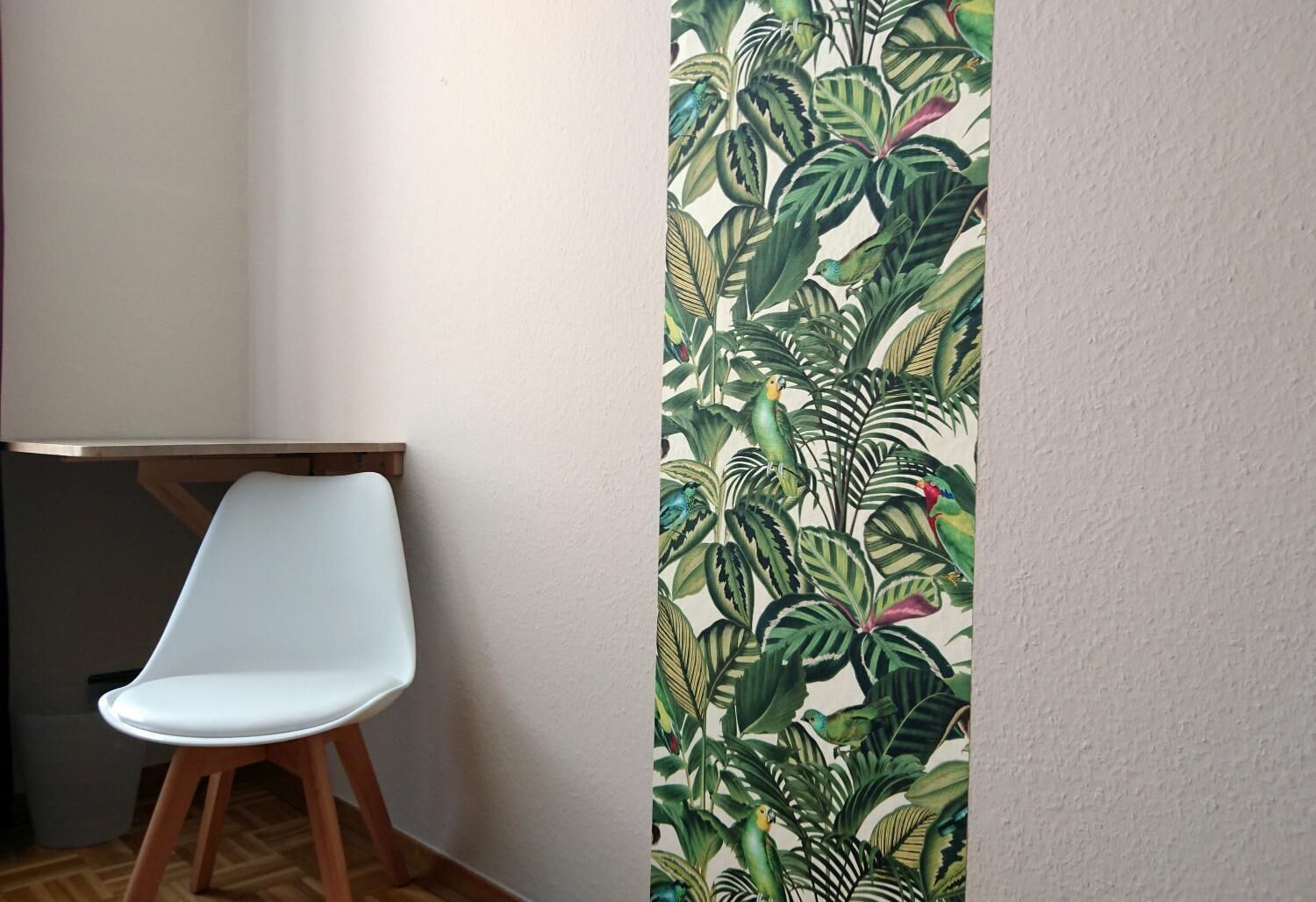 A corner of a room with a white chair, a wooden table, and a wall with a tropical plant wallpaper.
