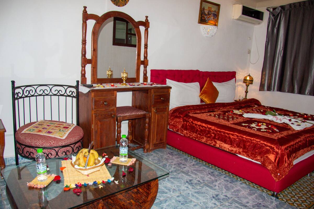 A bedroom with a bed, a wooden dresser, a glass table with a basket of fruit, and a chair.