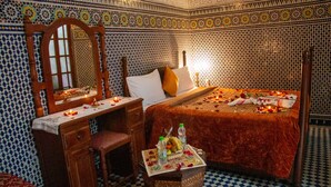 A room with a bed, a wooden desk, and a chair. The bed has a red blanket and pillows. There is a small table with a basket of fruit and bottles. The walls have a patterned tile design. - Riad Baba (Fes)