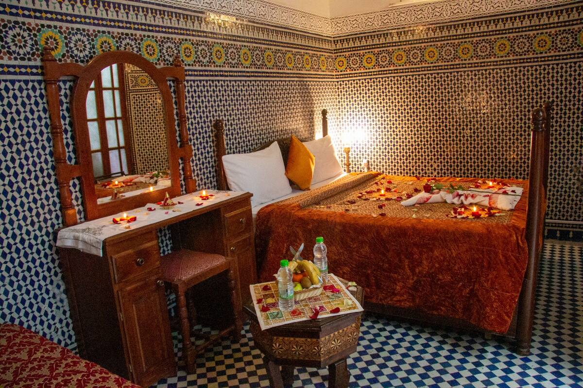 A room with a bed, a wooden desk, and a chair. The bed has a red blanket and pillows. There is a small table with a basket of fruit and bottles. The walls have a patterned tile design.