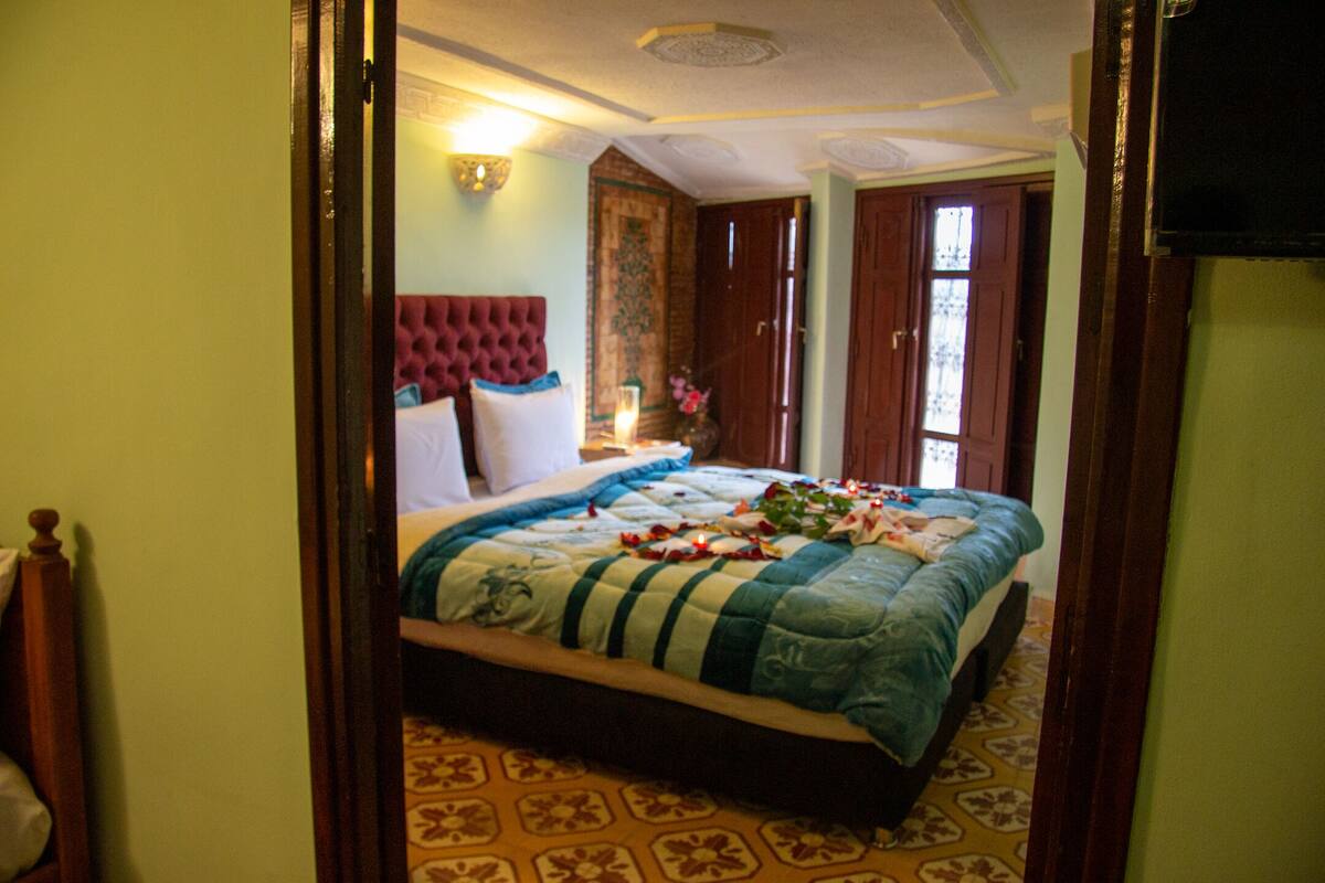 A bedroom with a large bed, a wooden headboard, a nightstand with a lamp, and a window with a view of the outside.