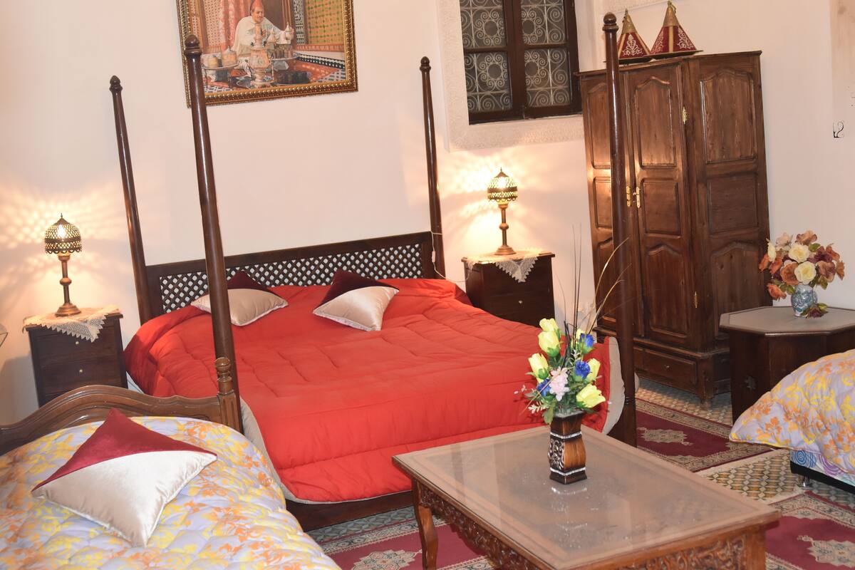 A four-poster bed with a red bedspread, a wooden wardrobe, a nightstand with a lamp, and a small table with a vase of flowers.