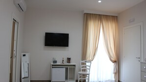 A room with a white desk, a television mounted on the wall, a white chair, a small refrigerator, and a door with a handle. - Philip Royal (Naples)