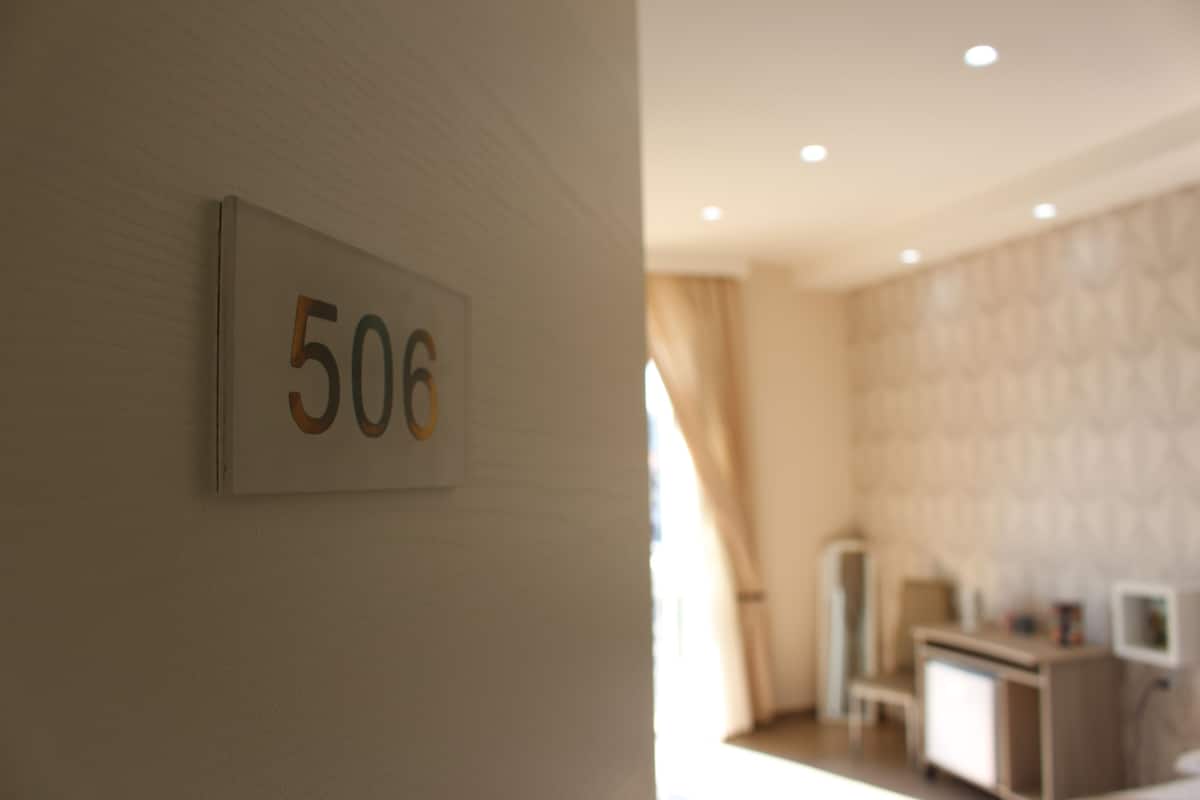 A room with a number sign '506' on the wall, a bed, a bedside table, and a television.