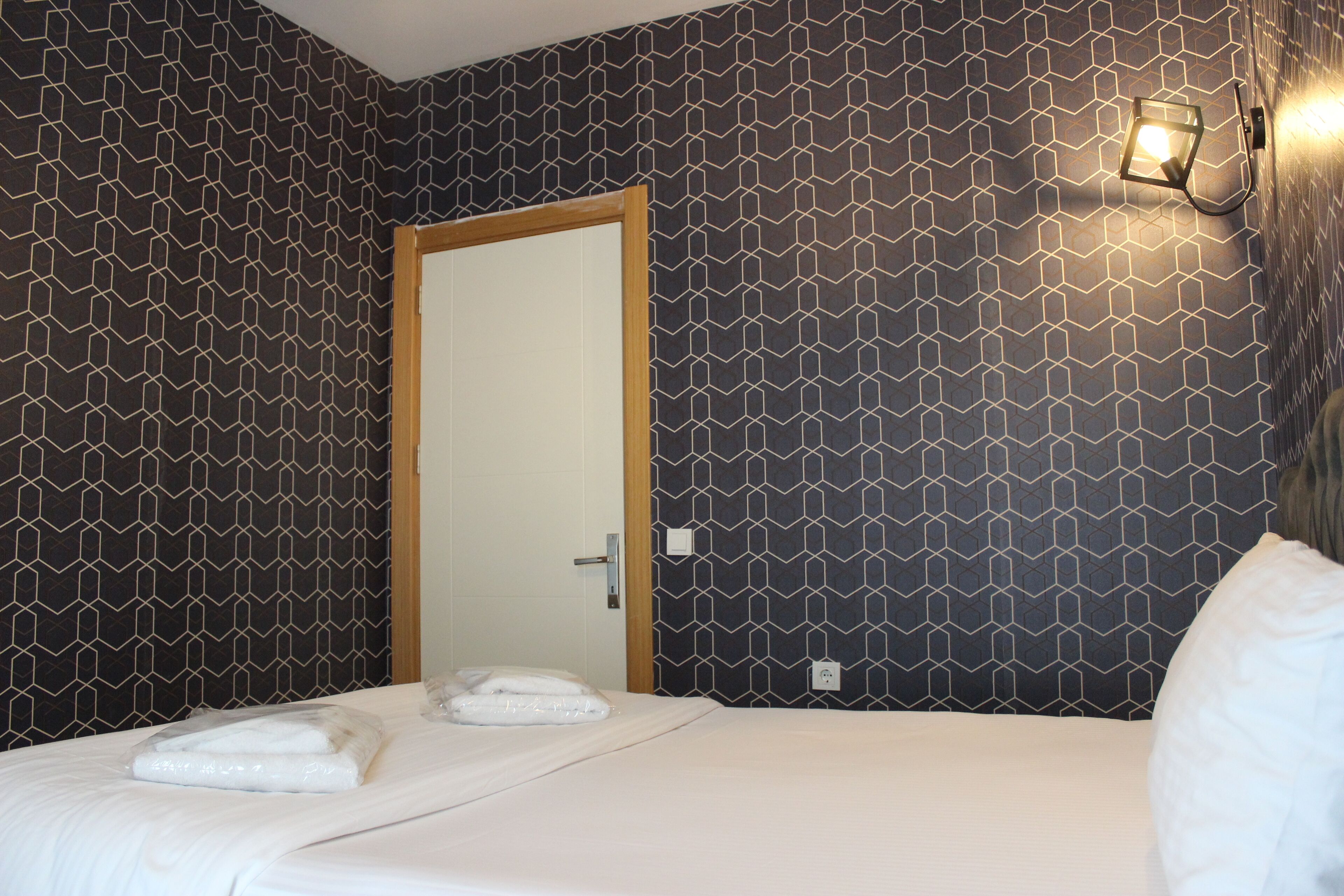 A hotel room with a geometric patterned wall, a wooden door, a bed with white linens, and a wall-mounted light fixture.