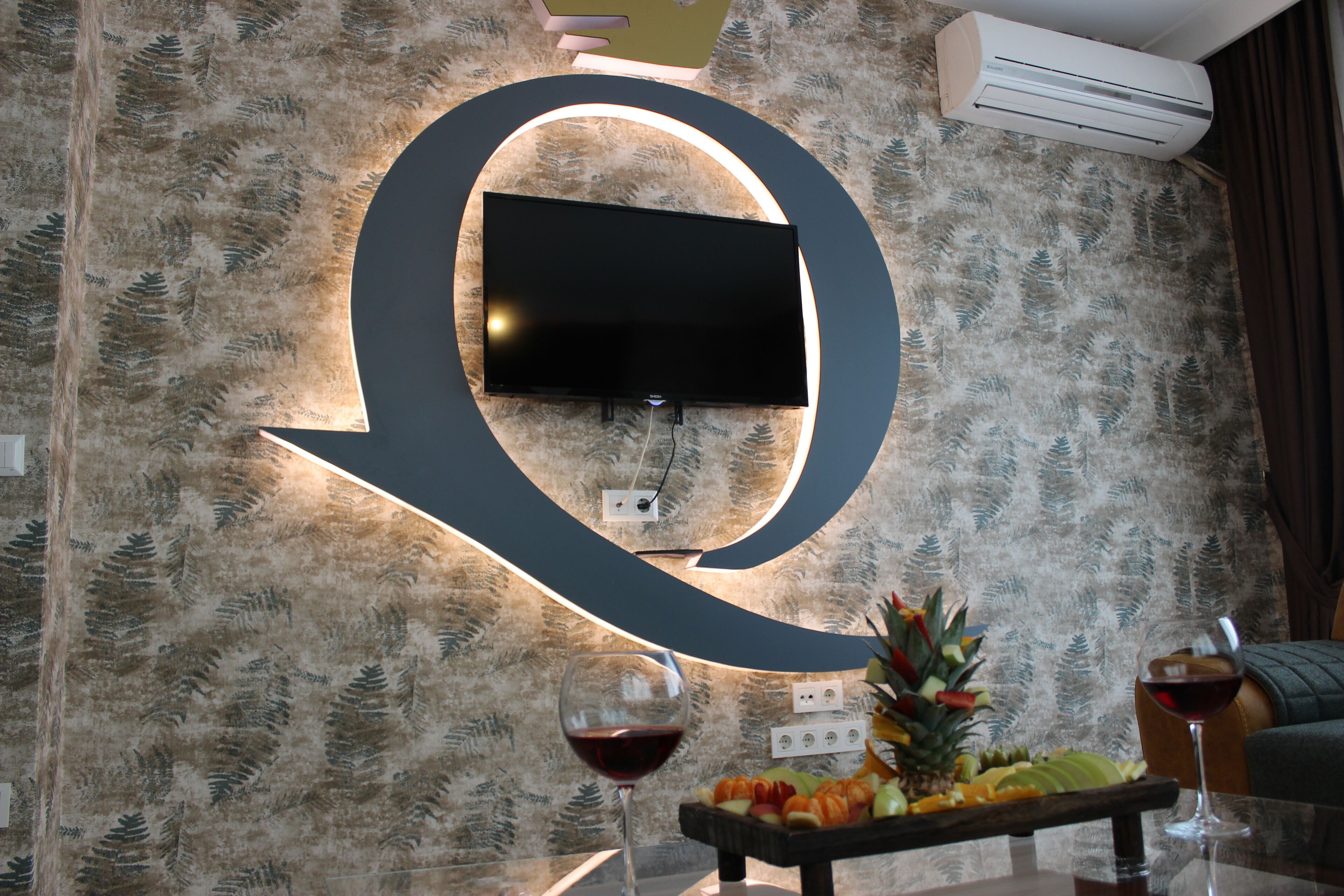Uma sala de estar moderna com um grande letreiro iluminado com a letra 'Q', uma TV de tela plana na parede e um prato de frutas sobre uma mesa de vidro.
