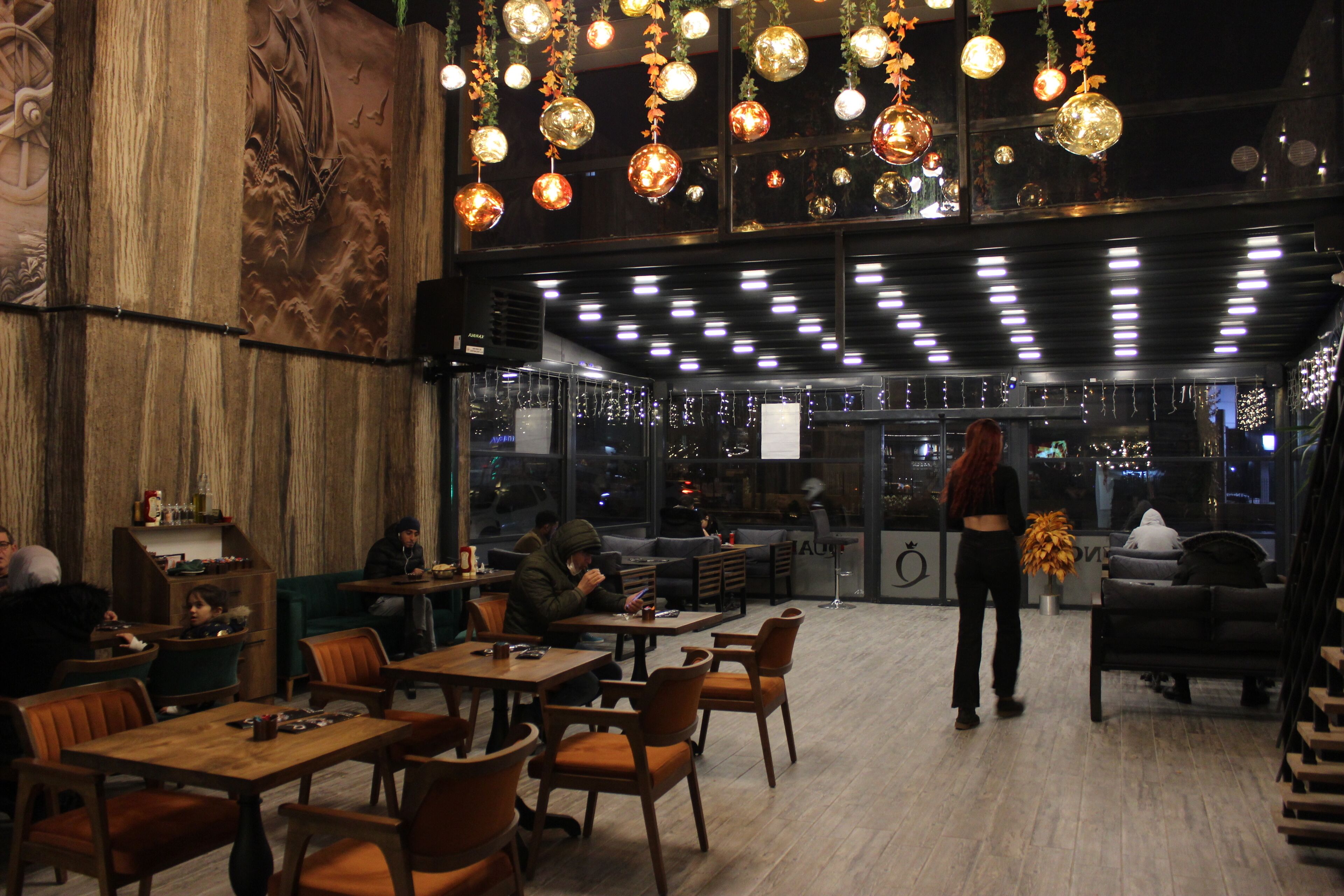 An indoor dining area with wooden tables and chairs, hanging spherical lights, and a large mural on the wall.