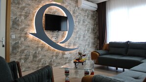 A modern living room with a large illuminated 'Q' on the wall, a flat-screen TV, a sofa, and a glass coffee table with fruit and wine glasses. - Quars Suite (Istanbul)