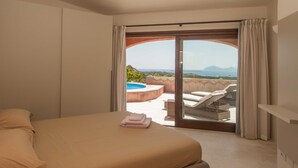 A bedroom with a bed, a view of an outdoor swimming pool, and a balcony with a chair and table. - Villa Pevero Hills 4 (Arzachena)