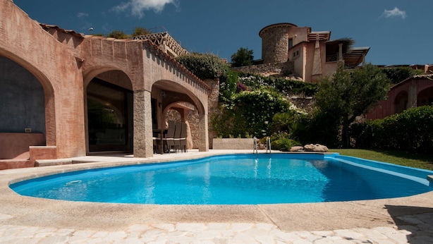 A swimming pool with stone tiles, surrounded by arched structures and a castle-like building in the background. - Villa Pevero Hills 4 (Arzachena)
