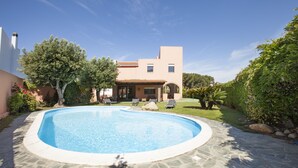 Una piscina con terraza de piedra, rodeada de vegetación y un edificio de dos plantas con tejado de baldosas.
