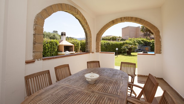 Un patio con una mesa y sillas de madera redonda, dos arcos de piedra y vistas a un jardín y edificios a lo lejos.