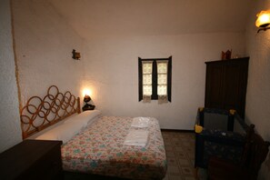 A bedroom with a bed, a nightstand, a window with curtains, a wooden headboard, a wooden cabinet, and a small table with a lamp. - Villa Scarabeo (Maracalagonis)