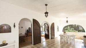 A spacious corridor with a tiled floor, wooden doors, and white walls. There is a dining area with a table and chairs, and a view of greenery through an arched window. - Villa Wanda (Quartu Sant'Elena)