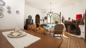 A dining area with a wooden table, chairs, and a plate of food. There is a television, a fireplace, and various decorative items on the walls. - Villa Wanda (Quartu Sant'Elena)