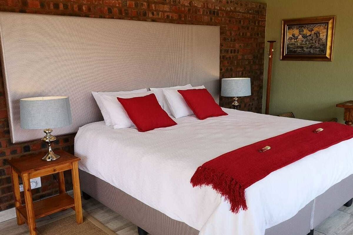 Una cama bien tendida con ropa blanca y almohadas rojas, una mesita de noche de madera con una lámpara y un muro de ladrillo de fondo.