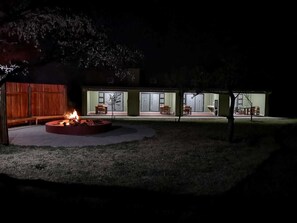 Vue nocturne d’une propriété résidentielle avec un foyer extérieur, un espace salon extérieur et une terrasse couverte.