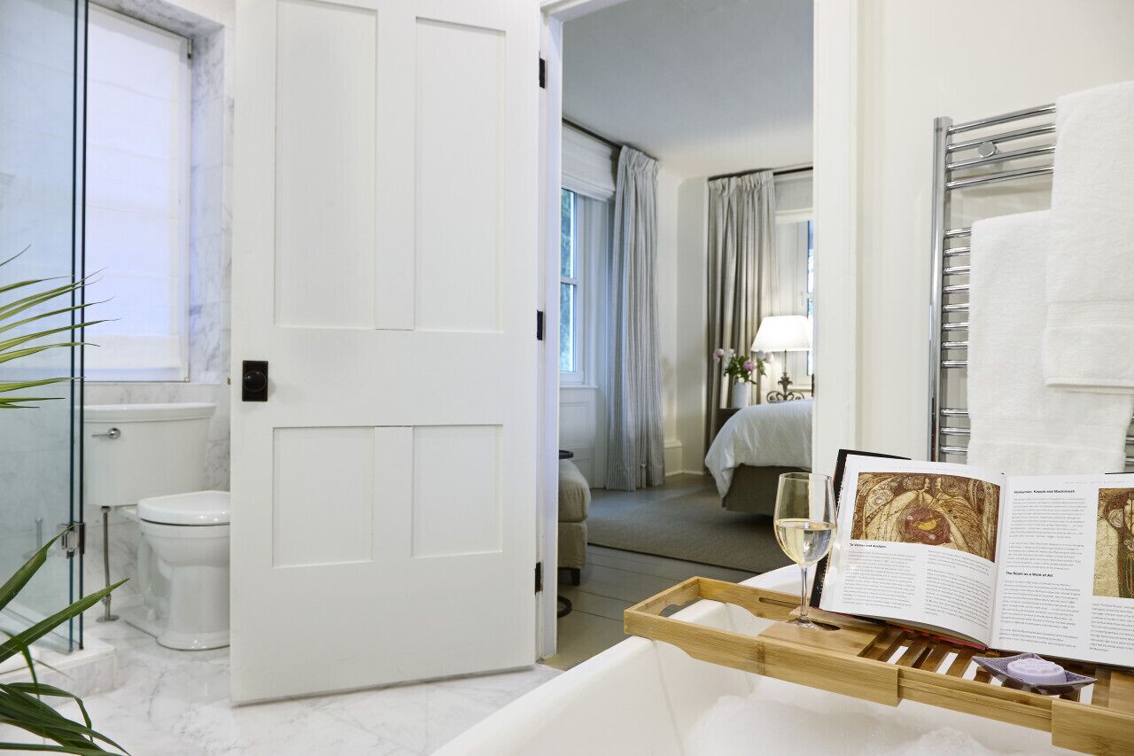 Quarto de hotel com banheiro, cama, mesa de cabeceira com um livro e um copo de vinho, e vista para o corredor.