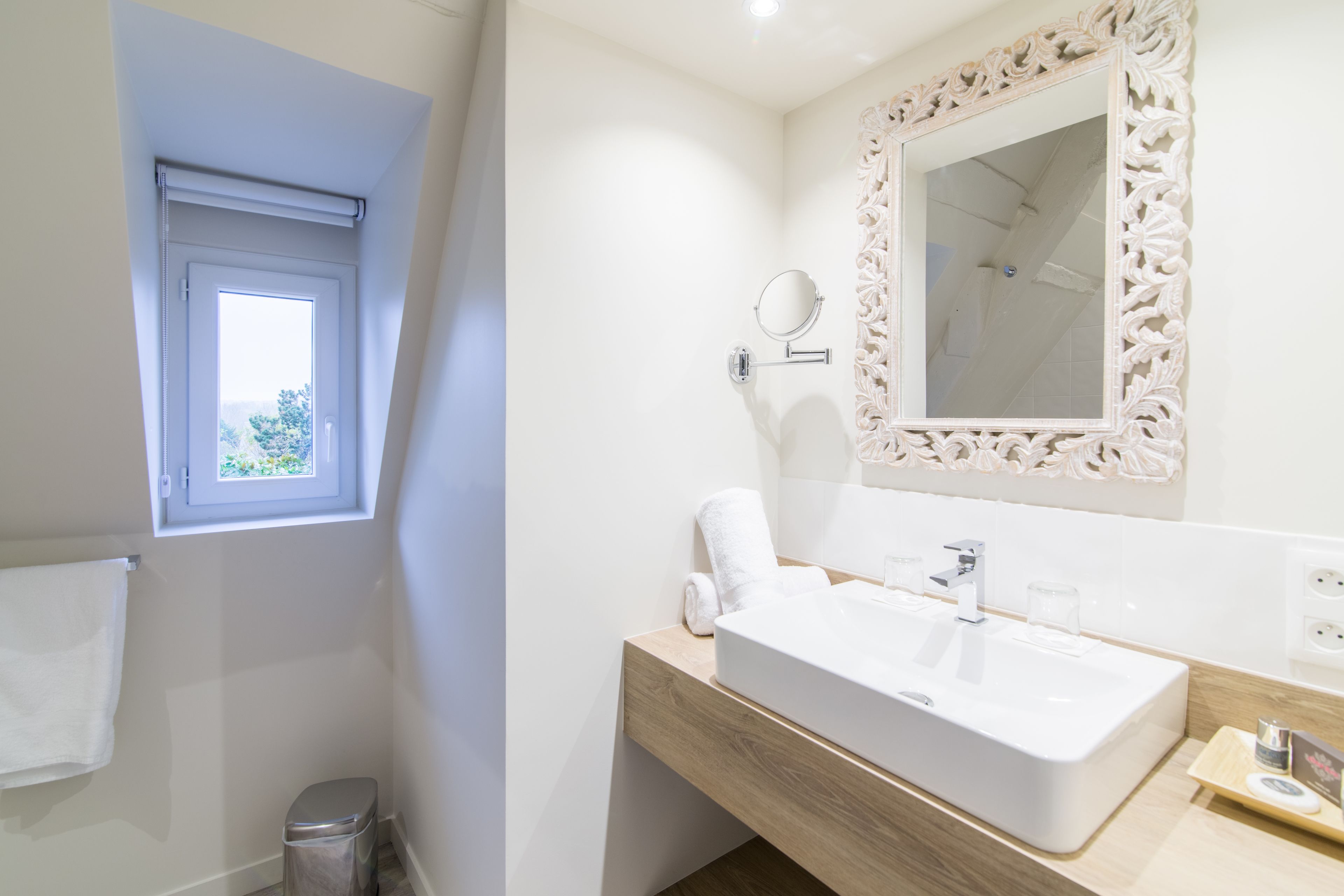 A modern bathroom with a large white sink, a mirror with an ornate frame, and a window with a view of greenery.