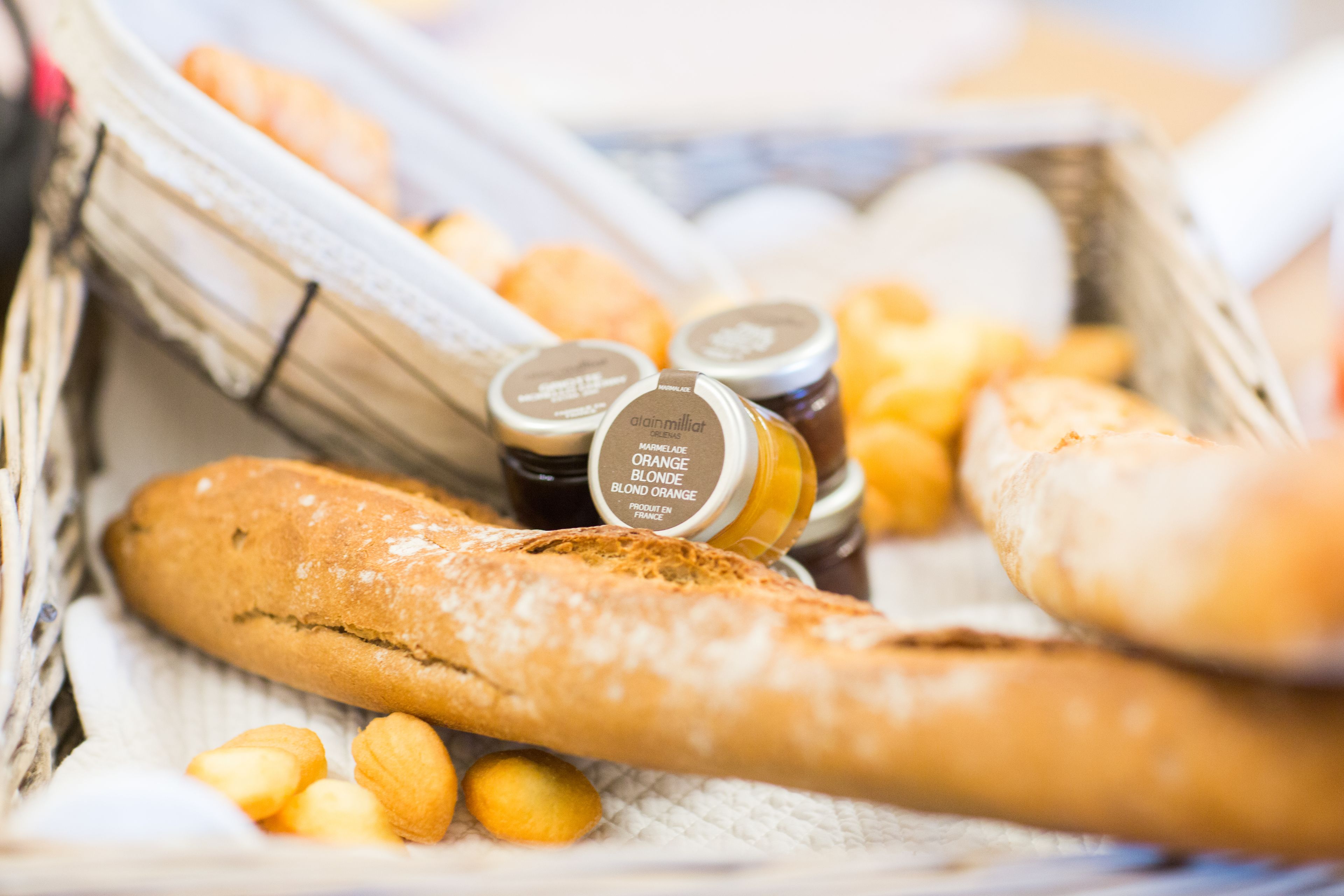 Una cesta con una baguette, pequeños pasteles y frascos de miel naranja y rubia.