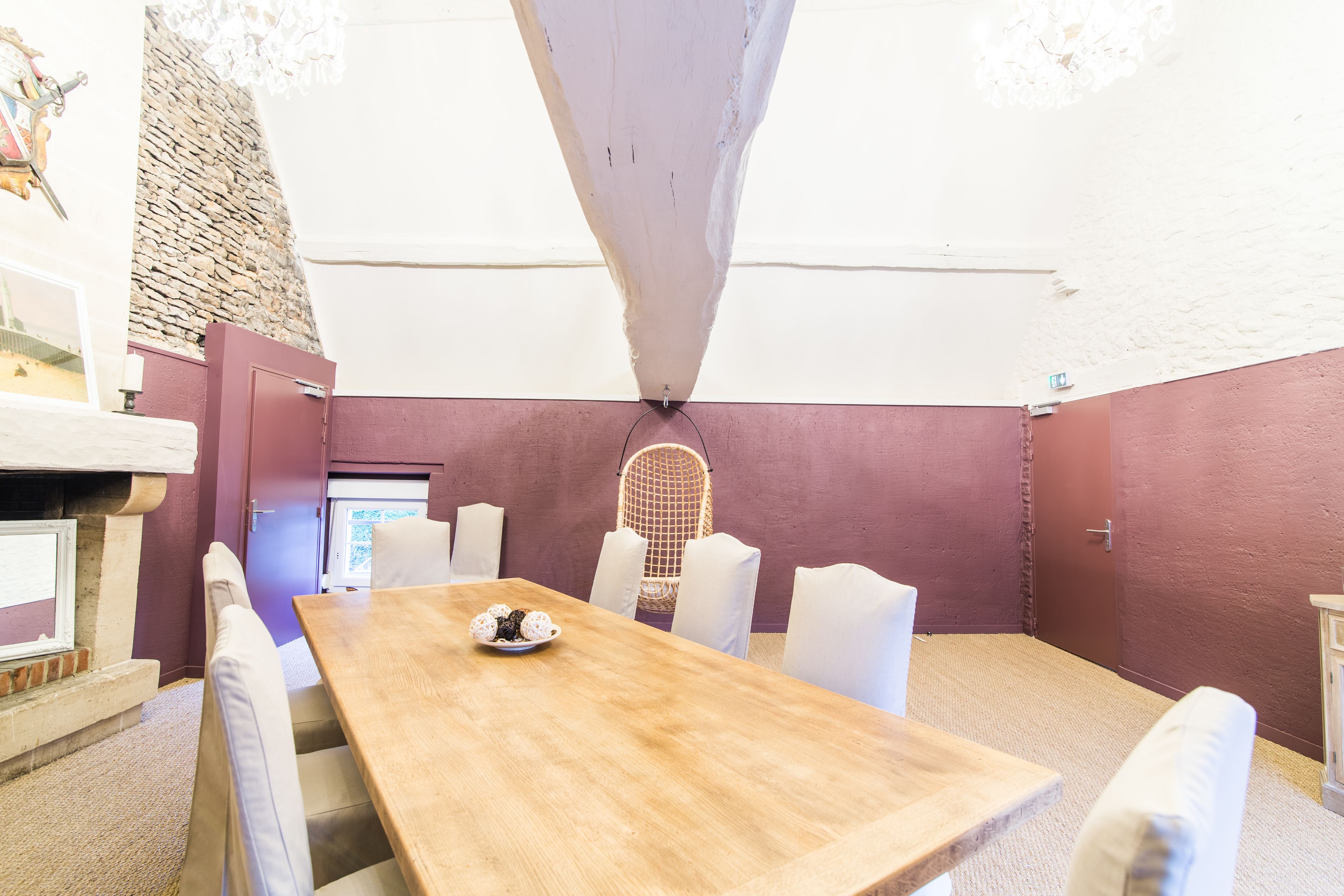 Un espace repas comprenant une table en bois, des chaises blanches, un fauteuil en osier et un mur en pierre.
