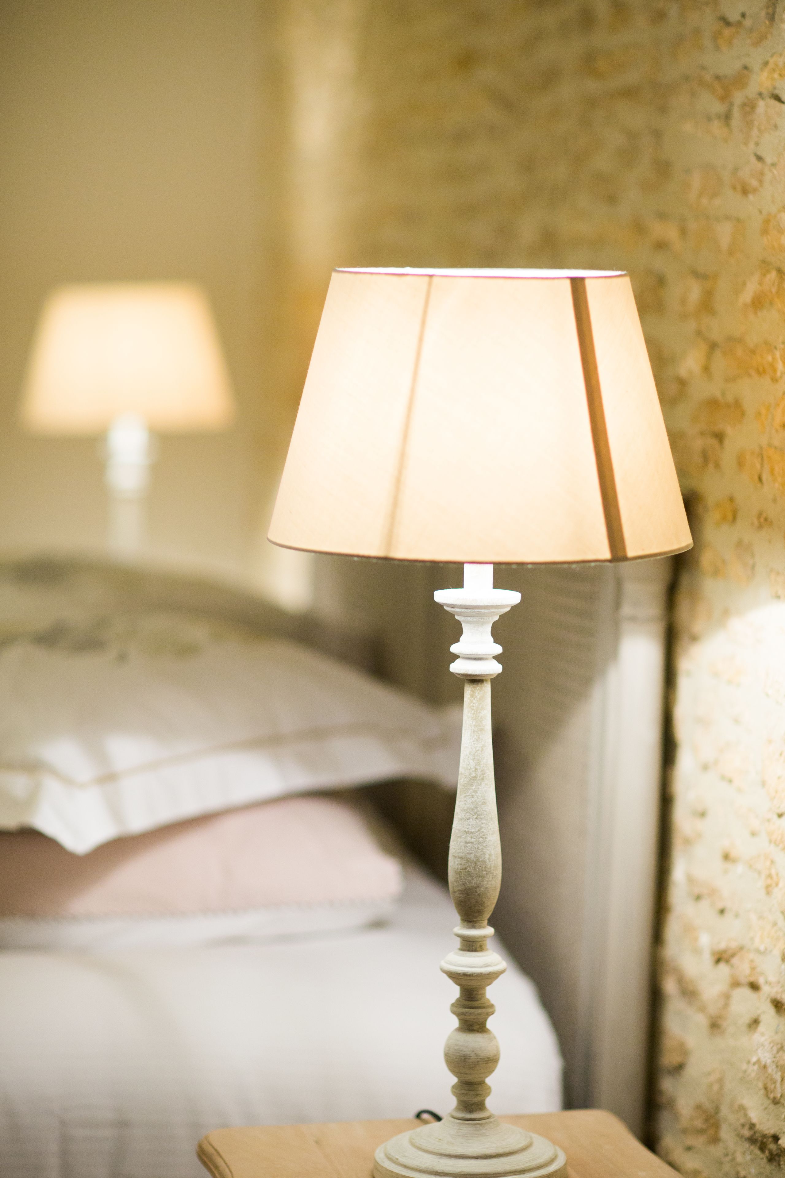 A bedside table with a lamp and a bed with pillows in the background.