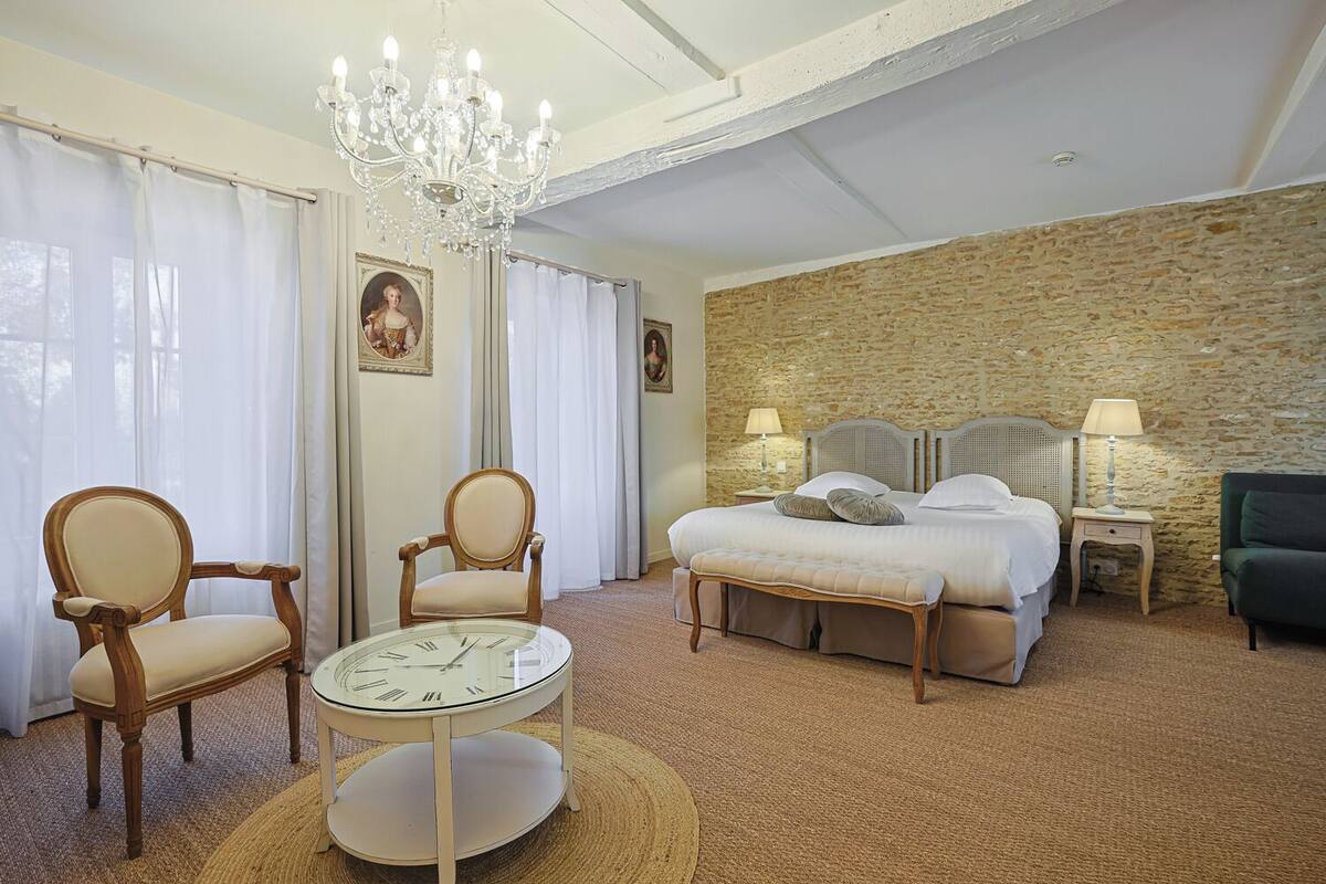 Een hotelkamer met een groot bed, twee fauteuils, een ronde tafel met een klok, een kroonluchter en een stenen muur.