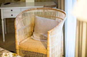 Una silla de mimbre con cojín, un escritorio blanco y una ventana con cortinas.