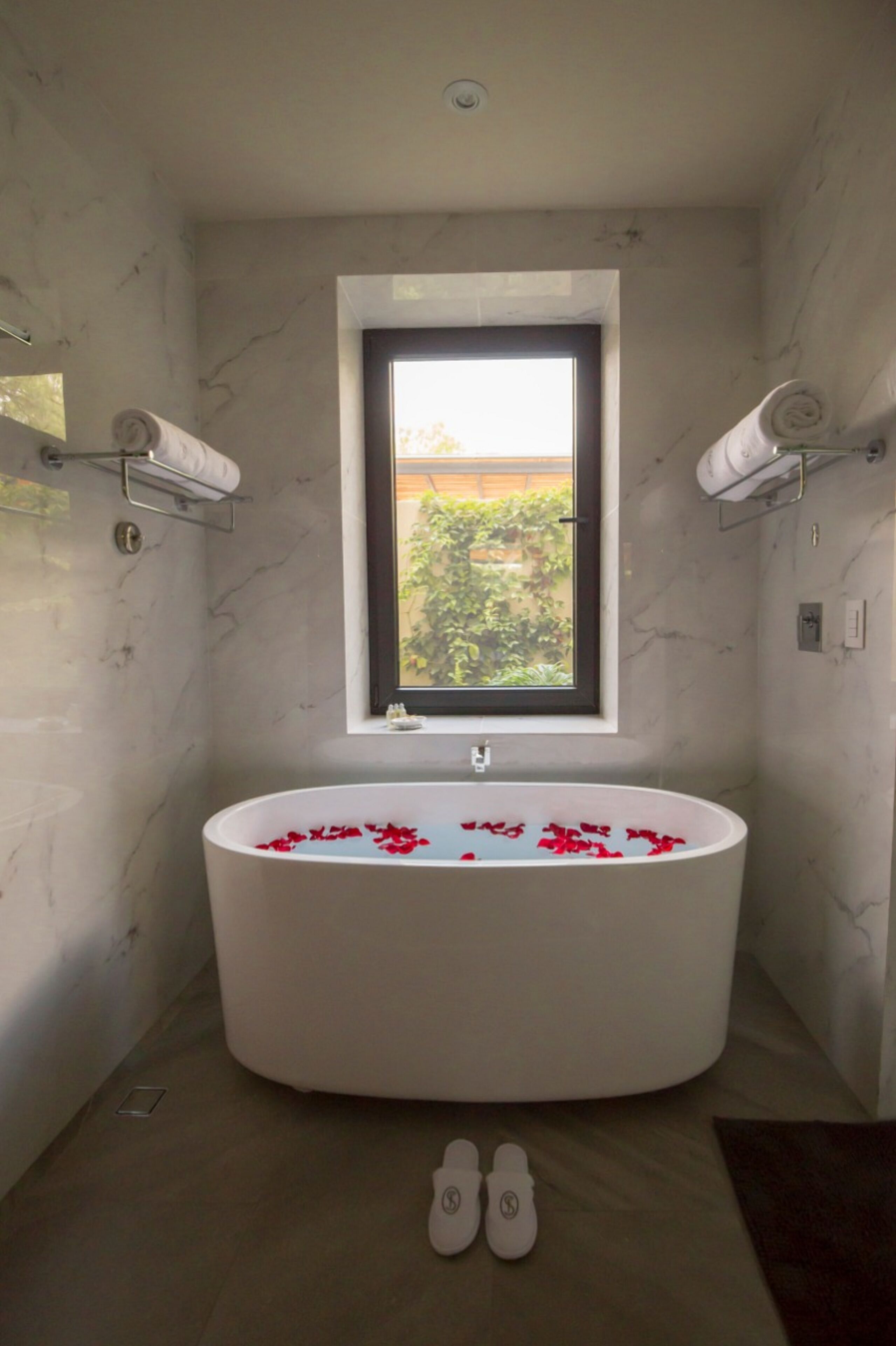 A bathroom with a freestanding bathtub filled with rose petals, a window above the tub, and a pair of slippers on the floor.