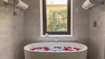 A bathroom with a freestanding bathtub filled with rose petals, a window above the tub, and a pair of slippers on the floor.