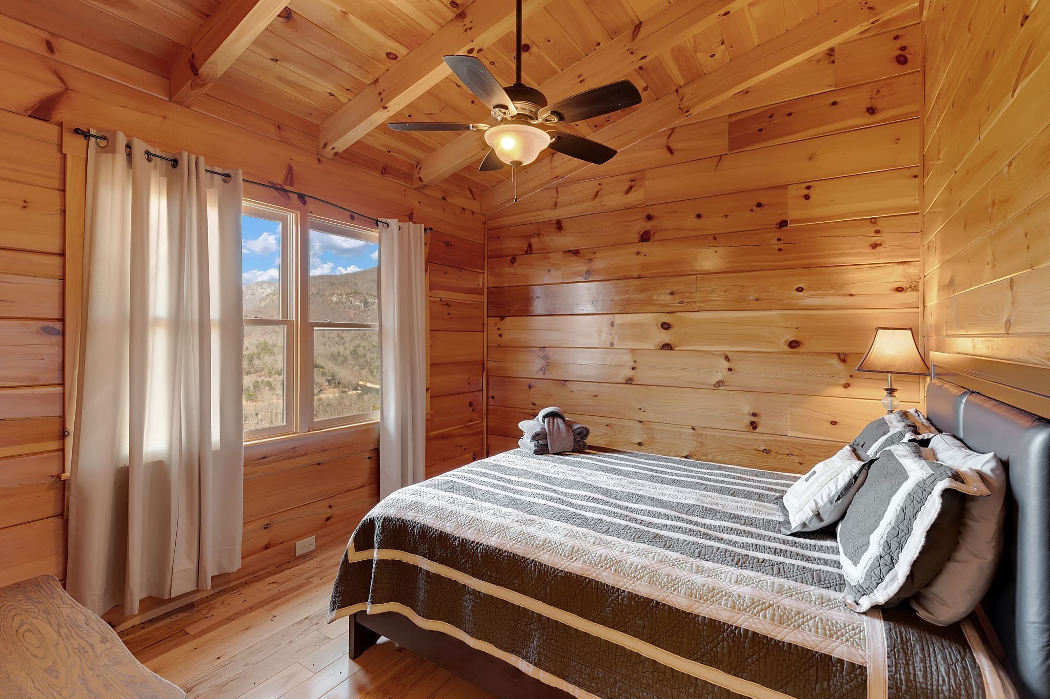 Un dormitorio de cabaña de madera con cama, cortinas, ventilador de techo y ventana con vistas a las montañas.