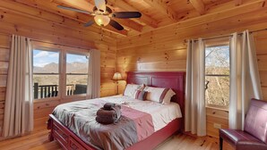 Quarto de cabana de madeira com cama, ventilador de teto e vista para montanhas pelas janelas.