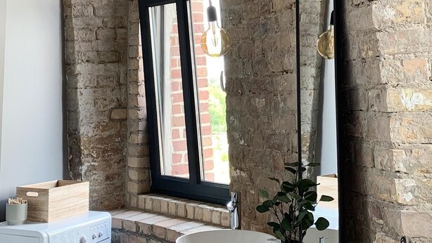 A rustic bathroom with exposed brick walls, a large arched window, a round sink, and a washing machine.