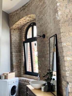 A rustic bathroom with exposed brick walls, a large arched window, a round sink, and a washing machine.