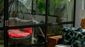 Ein Zimmer mit einem großen Fenster, einem Hängekorbstuhl mit roten Kissen, einem weißen Sofa mit gemusterten Kissen und einem Holznachttisch.
