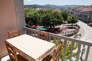 A balcony with a checkered tablecloth, wooden chairs, and a view of a street with trees and buildings. - Lethes View Apartamentos Turisticos (Ponte da Barca)
