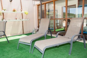 Two lounge chairs on artificial grass, a shelf with flowers, and a large window with a view of greenery. - Lethes View Apartamentos Turisticos (Ponte da Barca)