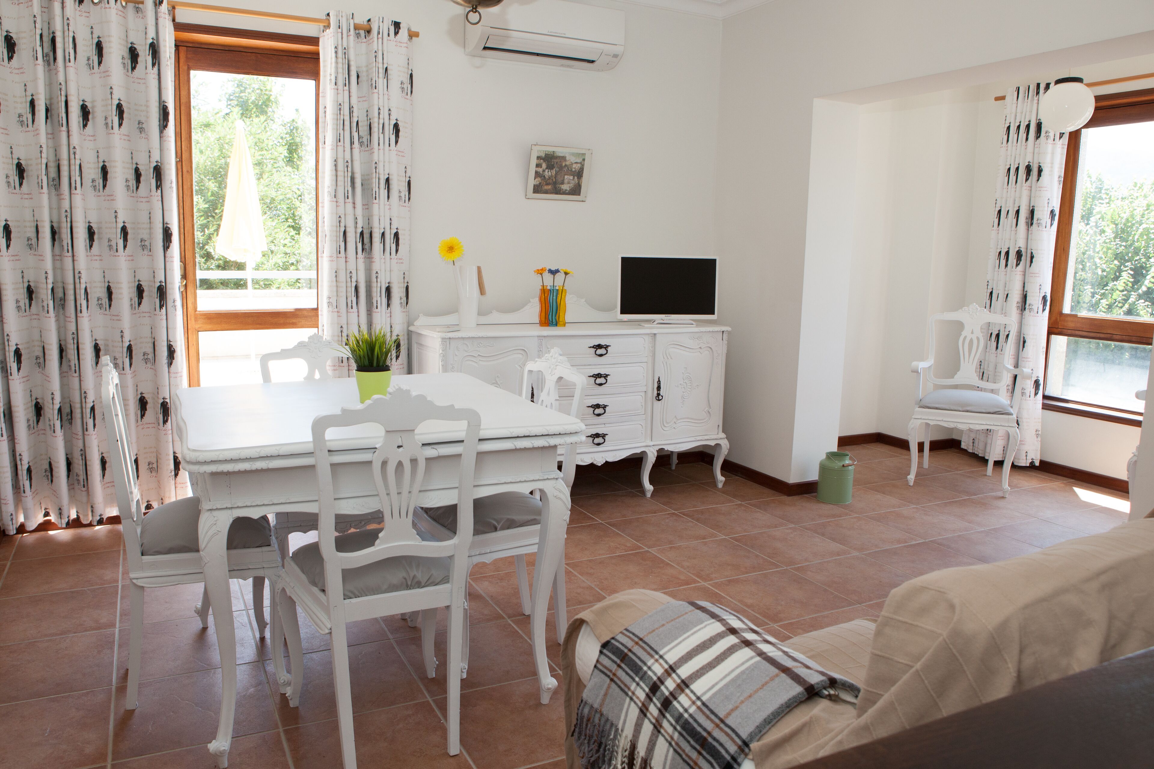 Una habitación con una mesa de comedor, sillas, un armario blanco, un televisor y una ventana con cortinas.