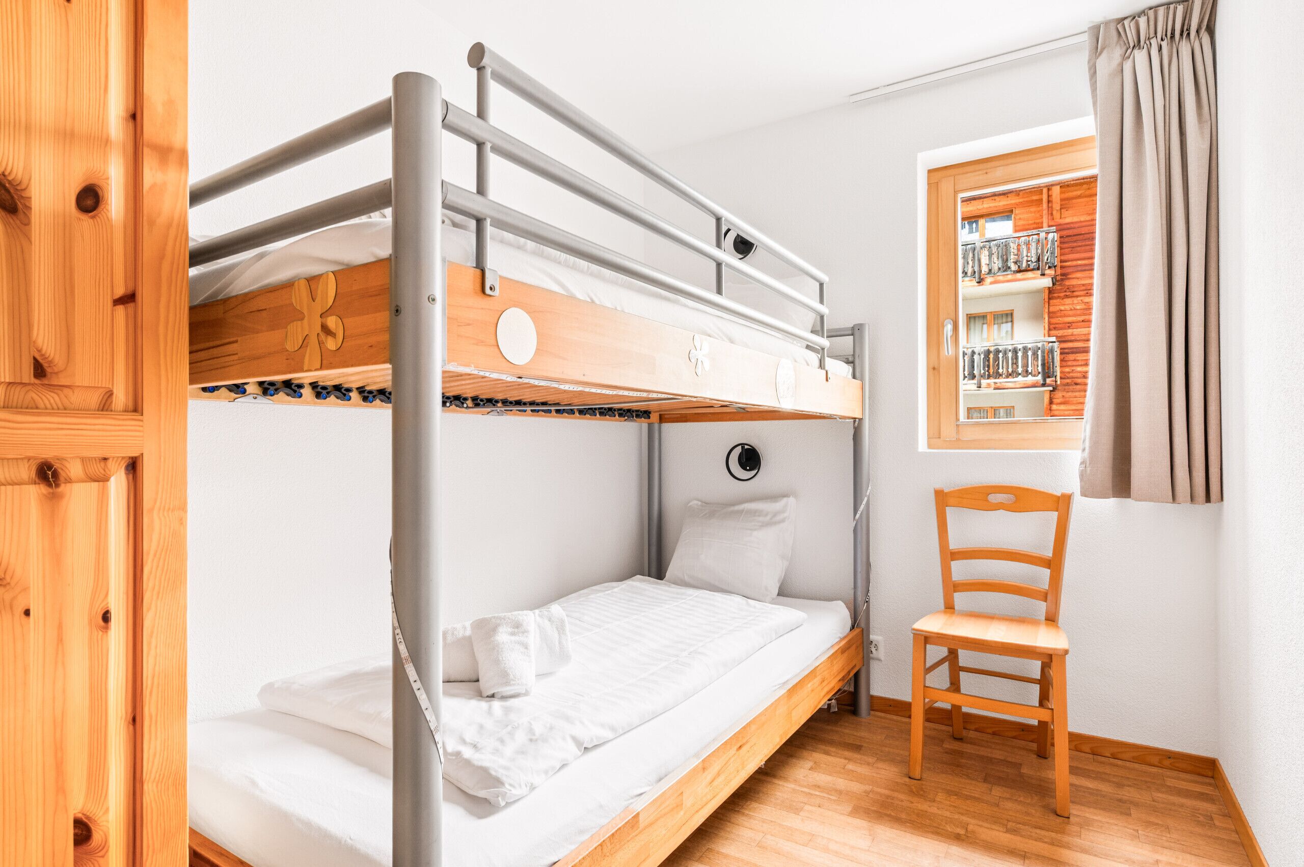 A bunk bed with a wooden headboard, a single bed below, a wooden chair, and a window with a view of a balcony.