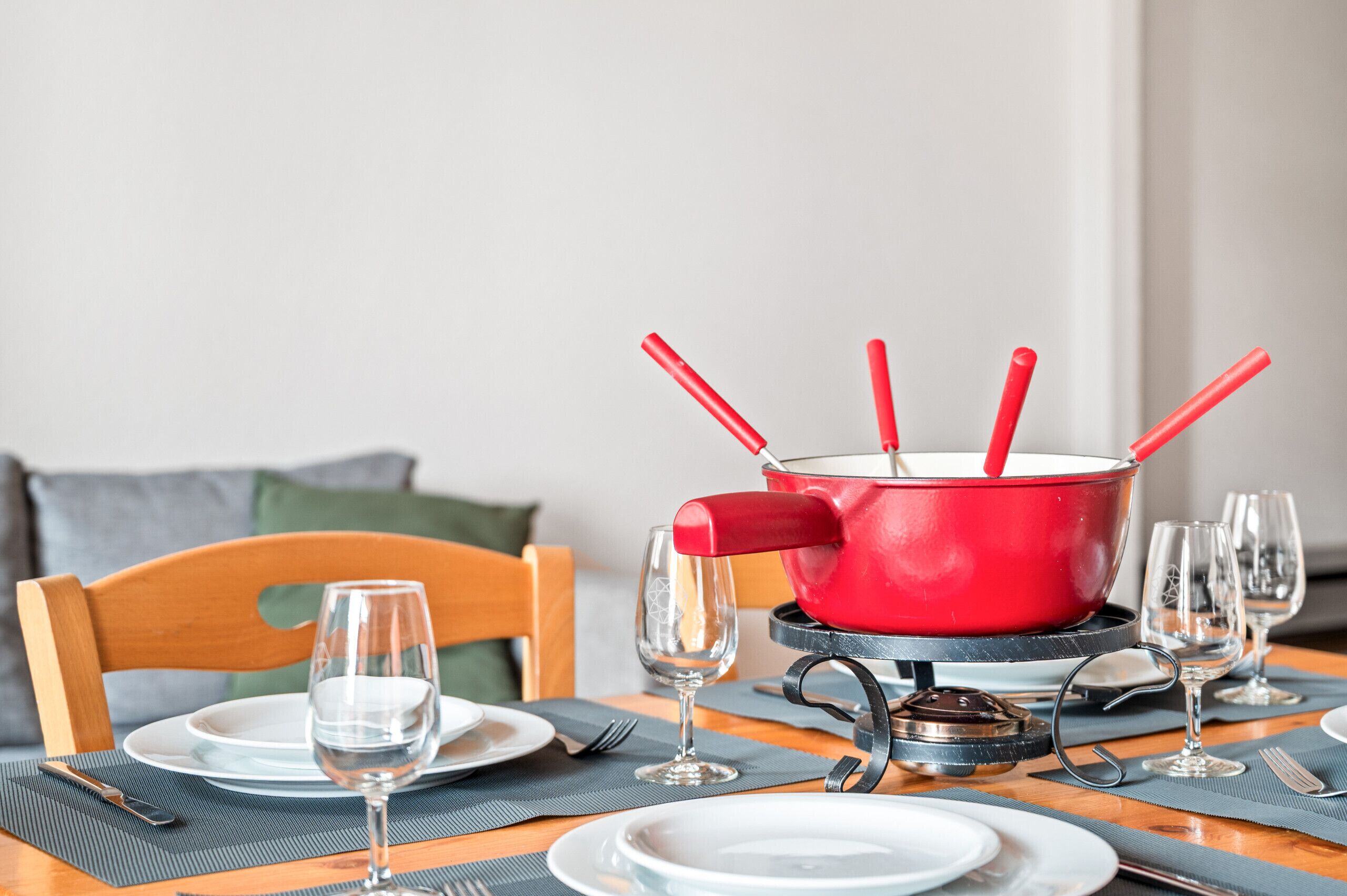 A dining table set with plates, cutlery, and wine glasses, with a red pot and spoons on a stand.