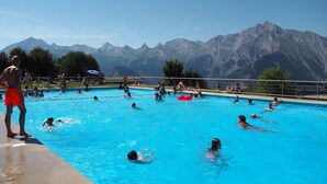 A swimming pool with people swimming and standing by the edge, surrounded by mountains.