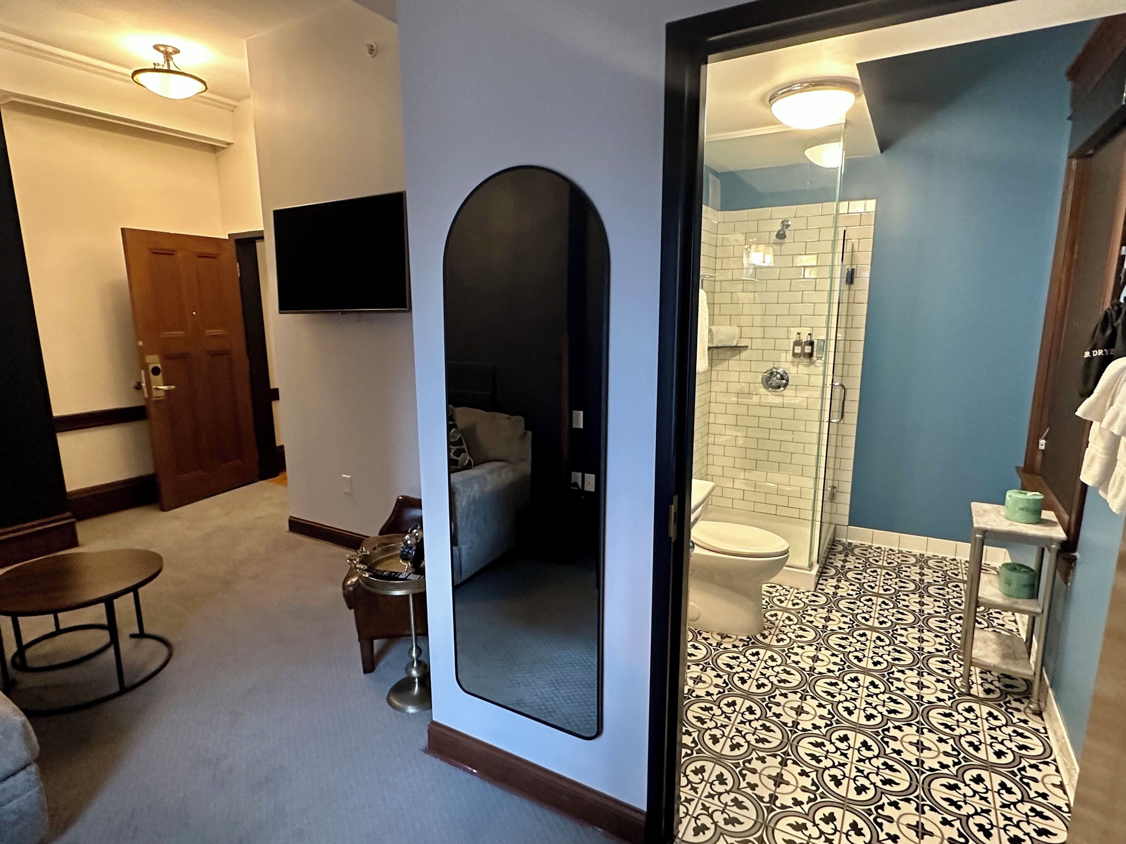A hotel room with a living area, a small table, a sofa, a television, and a bathroom with a shower, toilet, and patterned floor.