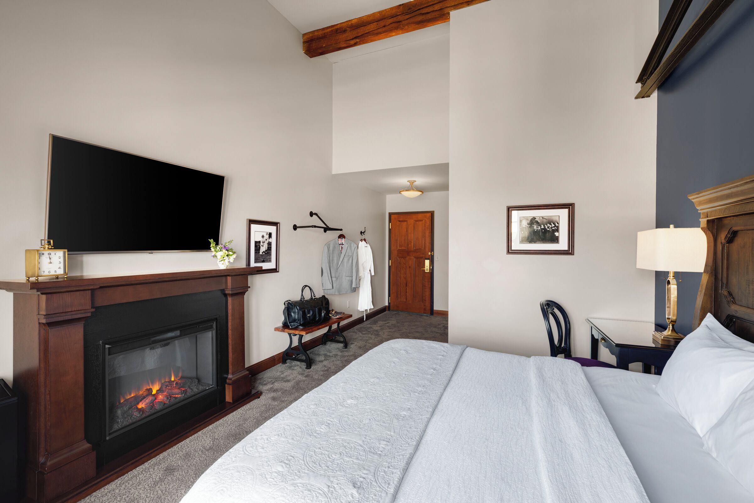 Habitación de hotel con una cama grande, chimenea, televisor de pantalla plana, escritorio con lámpara y puerta que da a otra habitación.