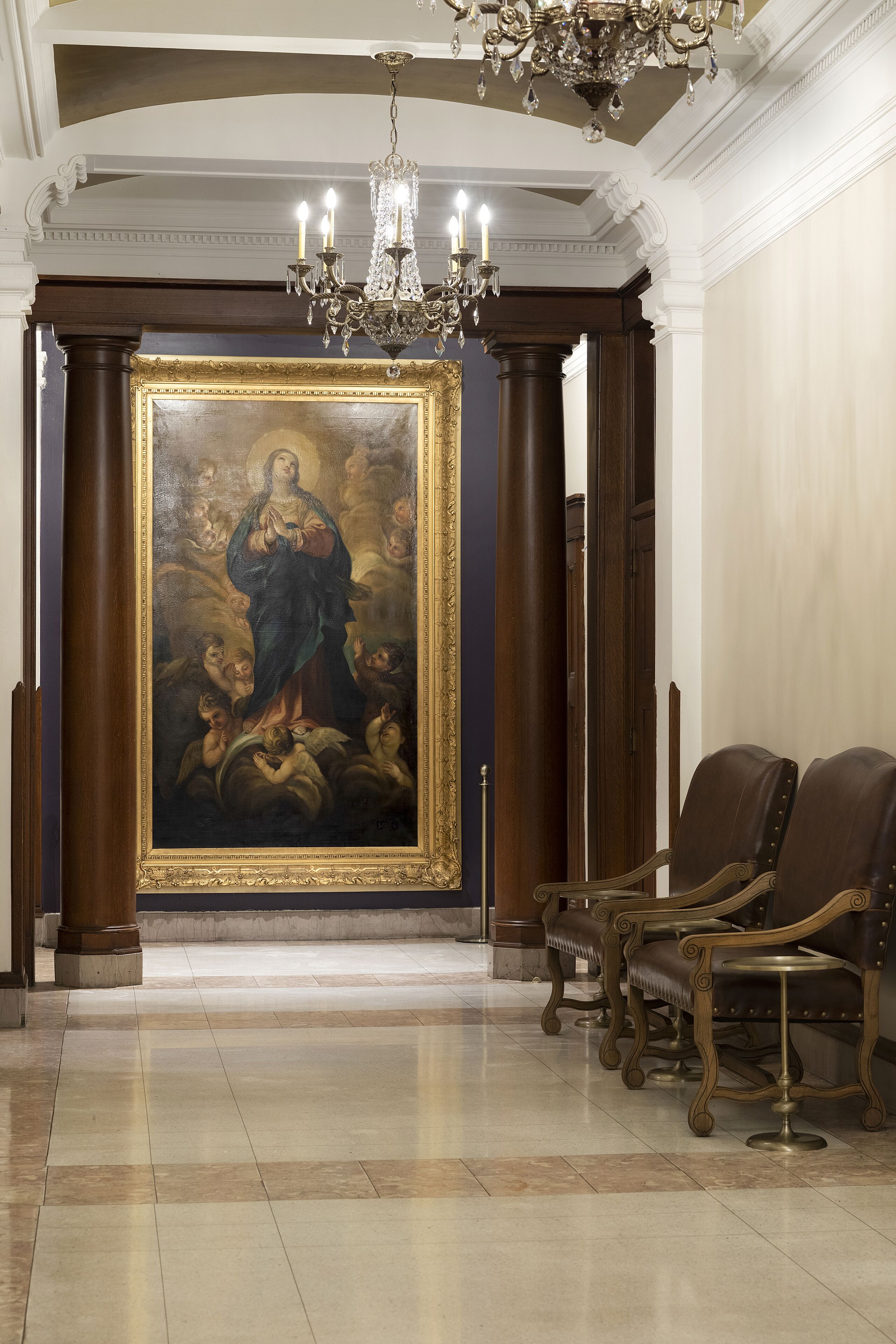 A grand hallway with a large religious painting, ornate columns, and a chandelier.