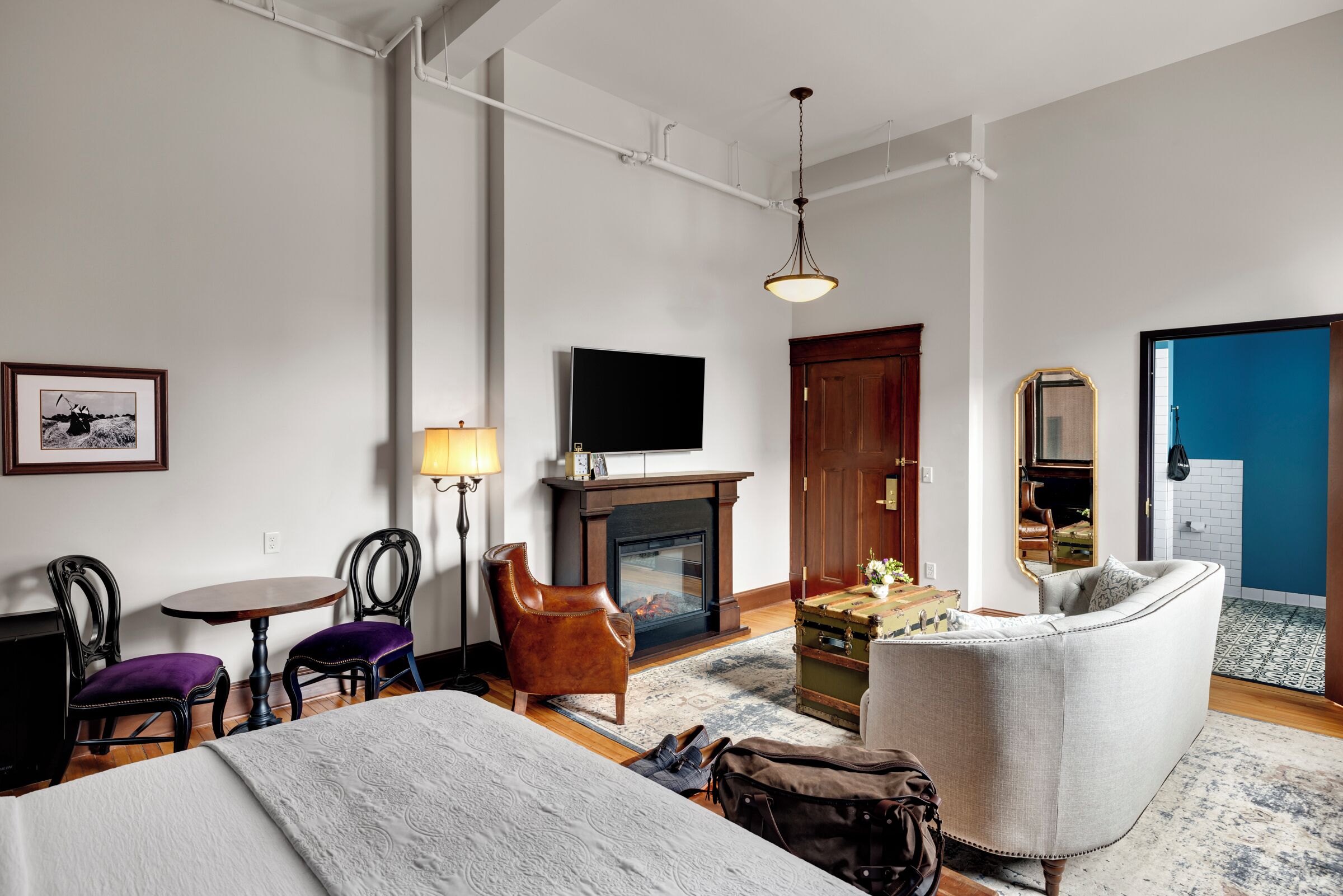 A hotel room with a bed, a chair, a table, a lamp, a fireplace, a television, and a suitcase.