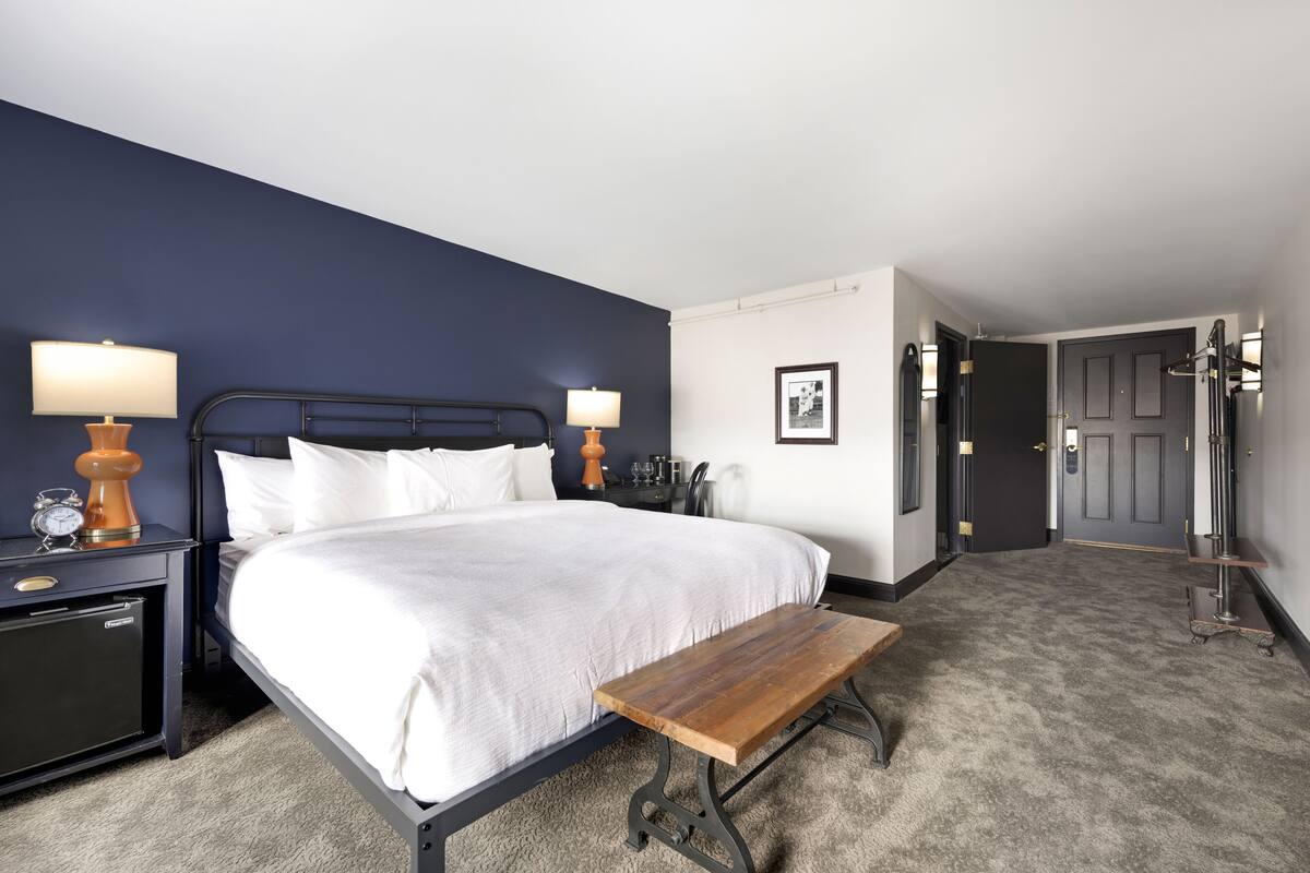 A hotel room with a bed, bedside tables, a bench, a nightstand, a lamp, a picture frame, and a door.