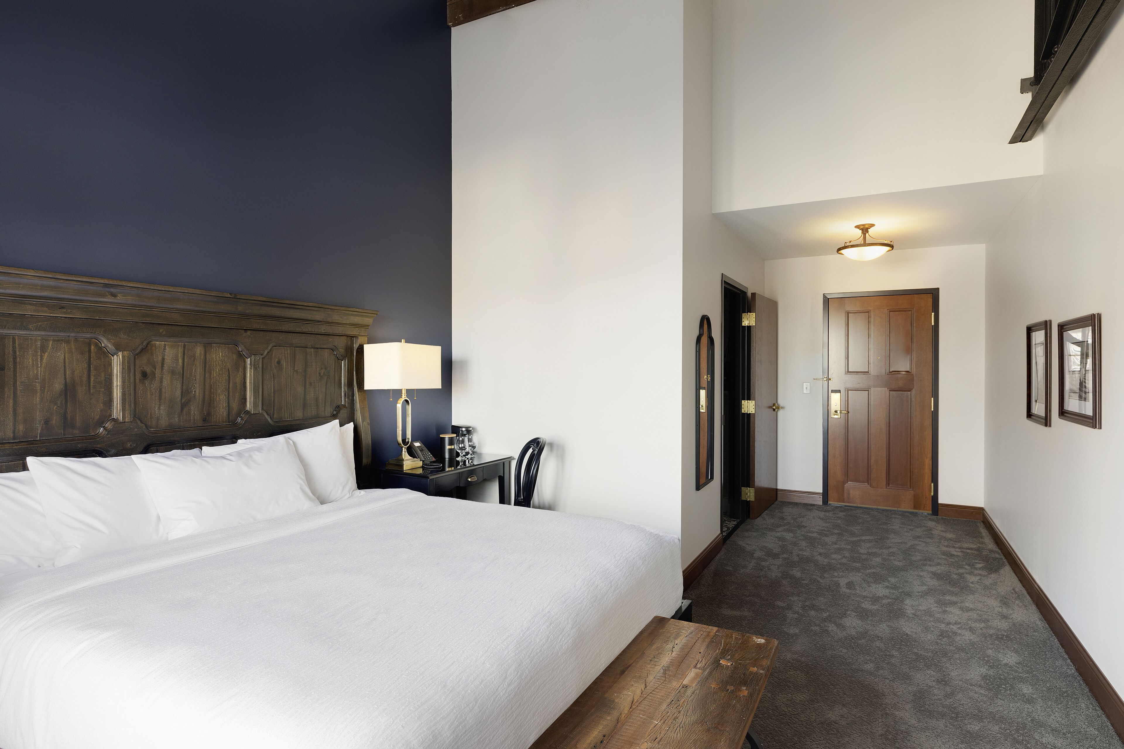 Habitación de hotel con una cama grande, cabecera de madera, mesita de noche con lámpara y escritorio con silla.