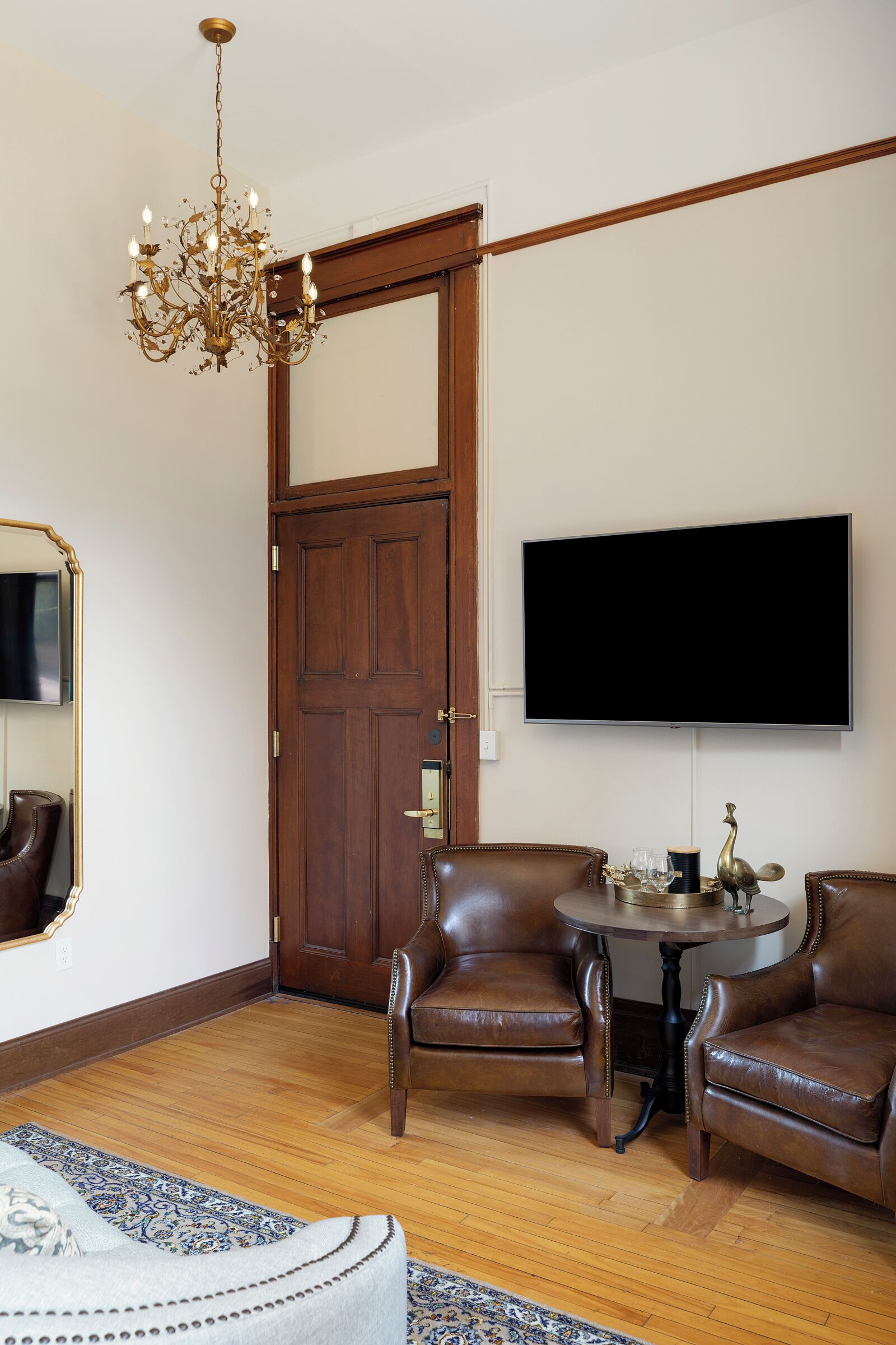 Une pièce dotée d’une porte en bois, d’un téléviseur fixé au mur, de deux fauteuils en cuir, d’une table ronde agrémentée d’objets décoratifs et d’un lustre.